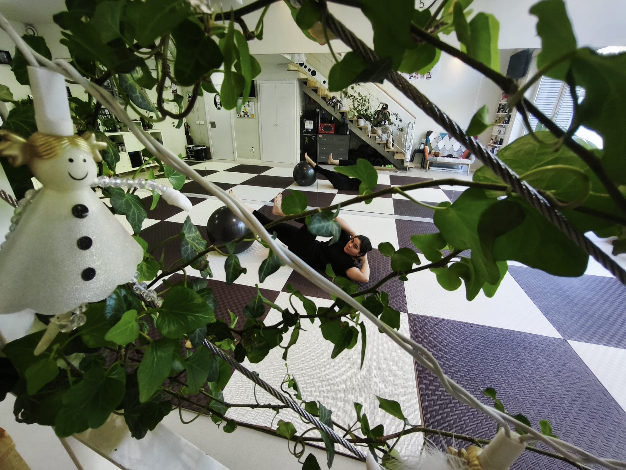 Ramas y hojas de una planta en primer plano y, detrás, una alumna tumbada sobre el suelo ajedrezado de la sala.